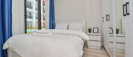 A modern bedroom with a large bed, white bedding, a nightstand, a wardrobe, and a view of a building outside.