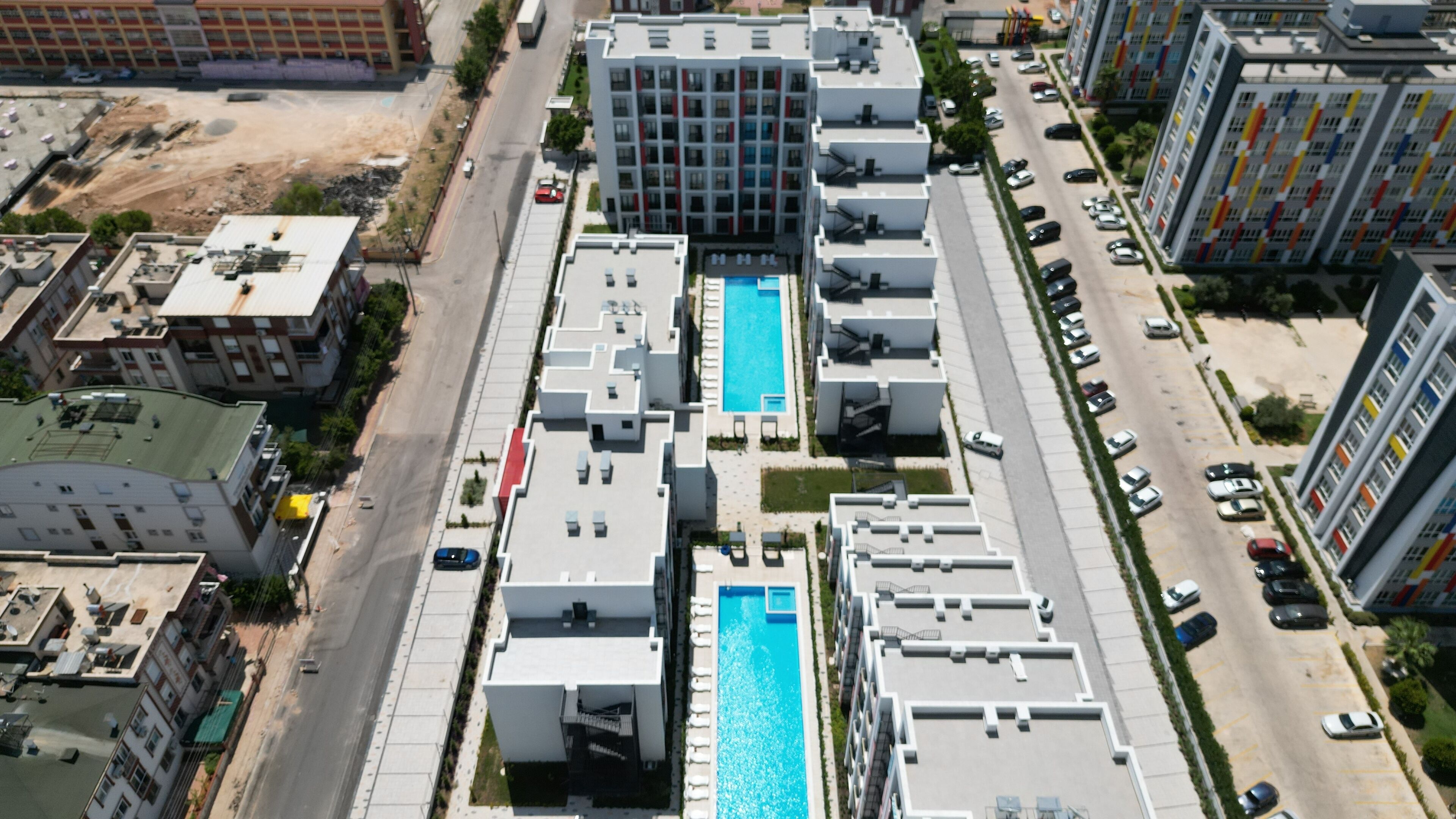 A modern apartment complex with a central swimming pool, multiple parking levels, and a mix of residential and commercial buildings in the vicinity.