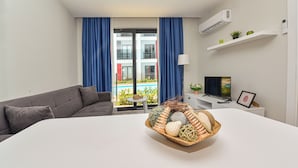 Una habitación de hotel moderna con un sofá gris, una mesa blanca, un televisor y vistas a una piscina a través de un ventanal.