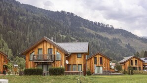 Eine Holzhütte mit Balkon, umgeben von einer grünen Wiese und einem bewaldeten Berg im Hintergrund.