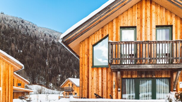Eine Holzhütte mit Balkon, umgeben von verschneitem Gelände und einem bewaldeten Berg im Hintergrund.