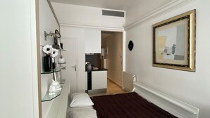 Um quarto com uma cama, um guarda-roupa branco, uma prateleira de vidro com artigos de higiene pessoal, um quadro na parede e uma cozinha ao fundo.