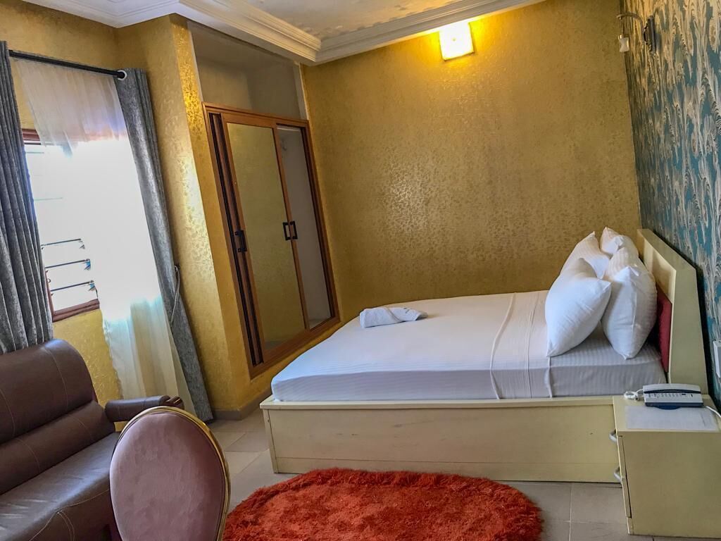 A hotel room with a bed, a red rug, a desk with a lamp, a wardrobe, and a window with curtains.