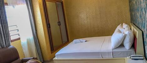 A hotel room with a bed, a red rug, a desk with a lamp, a wardrobe, and a window with curtains.