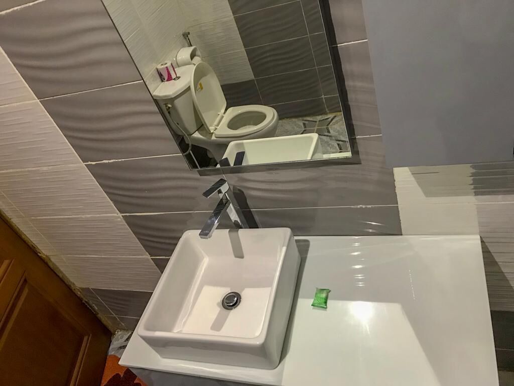 Une salle de bain avec un lavabo blanc, un miroir au-dessus, et des toilettes visibles dans le reflet.