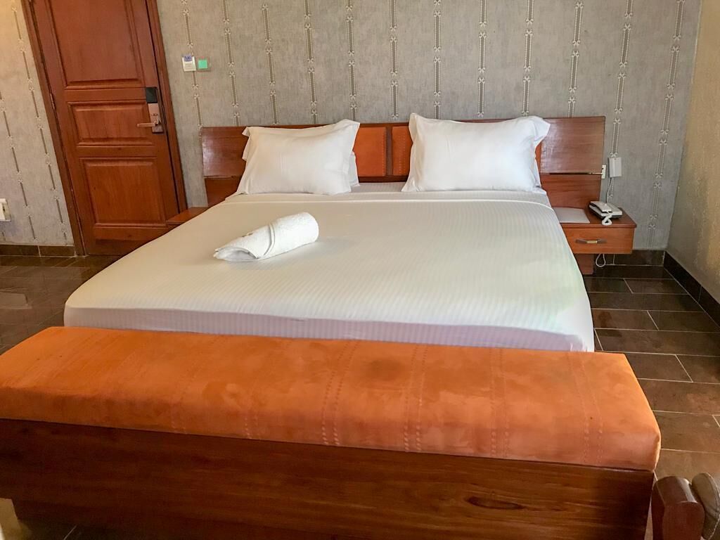 A neatly made bed with a white comforter and two pillows, a wooden nightstand with a lamp, and a wooden bench at the foot of the bed.