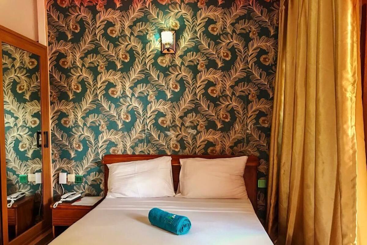 A hotel room with a bed, two pillows, a wooden headboard, a wall with floral wallpaper, a nightstand with a phone, and a curtain.