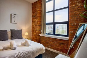A hotel room with a brick wall, a large window, a bed with white bedding, and a framed picture on the wall. - The Amber Suite (Manchester)