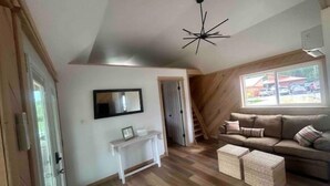 A spacious living area with a wooden floor, a white console table, a sofa with cushions, and a large window offering a view of the outside.