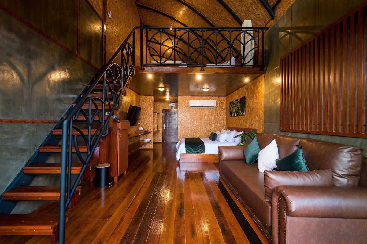 A wooden staircase with a metal railing leading to a loft bedroom with a bed and a brown leather sofa.