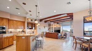 A spacious kitchen with wooden cabinets, a central island, and bar stools. There is a dining area with a round table and chairs, and a living room with a sofa and a fireplace. - Desert Oasis | TROON NORTH | Golf | Heated Pool (Scottsdale)