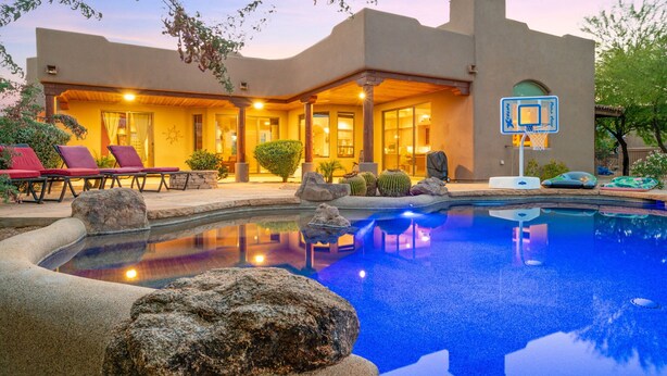 A pool with a basketball hoop, red lounge chairs, and a building with a patio. - Desert Oasis | TROON NORTH | Golf | Heated Pool (Scottsdale)