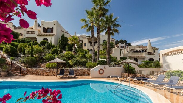 A swimming pool with lounge chairs, palm trees, and a Mediterranean-style building in the background. - La Manga Club Los Altos Villa 367 (Atamaría)
