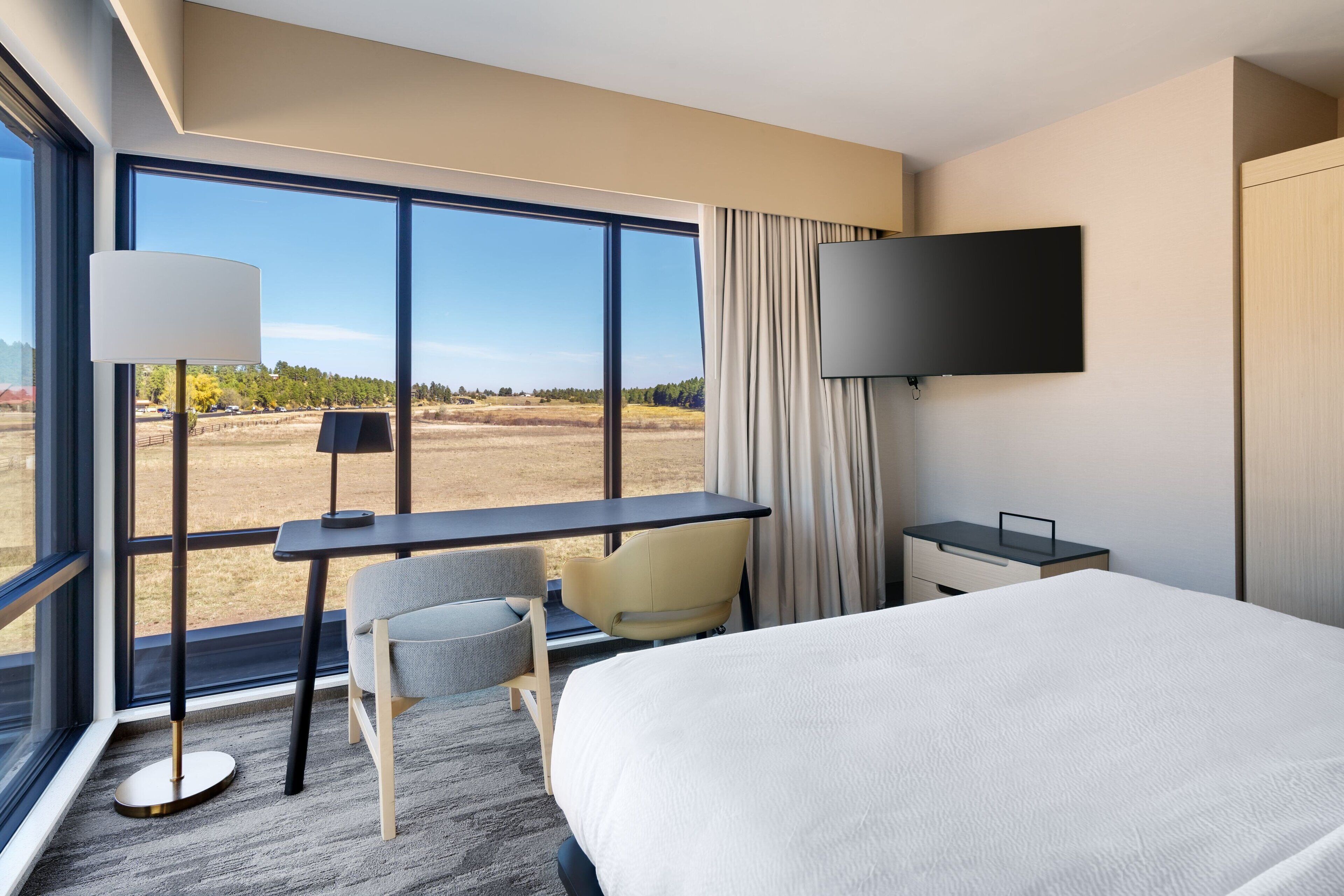 A hotel room with a large bed, a desk with a chair, a TV mounted on the wall, and a large window offering a view of a field.
