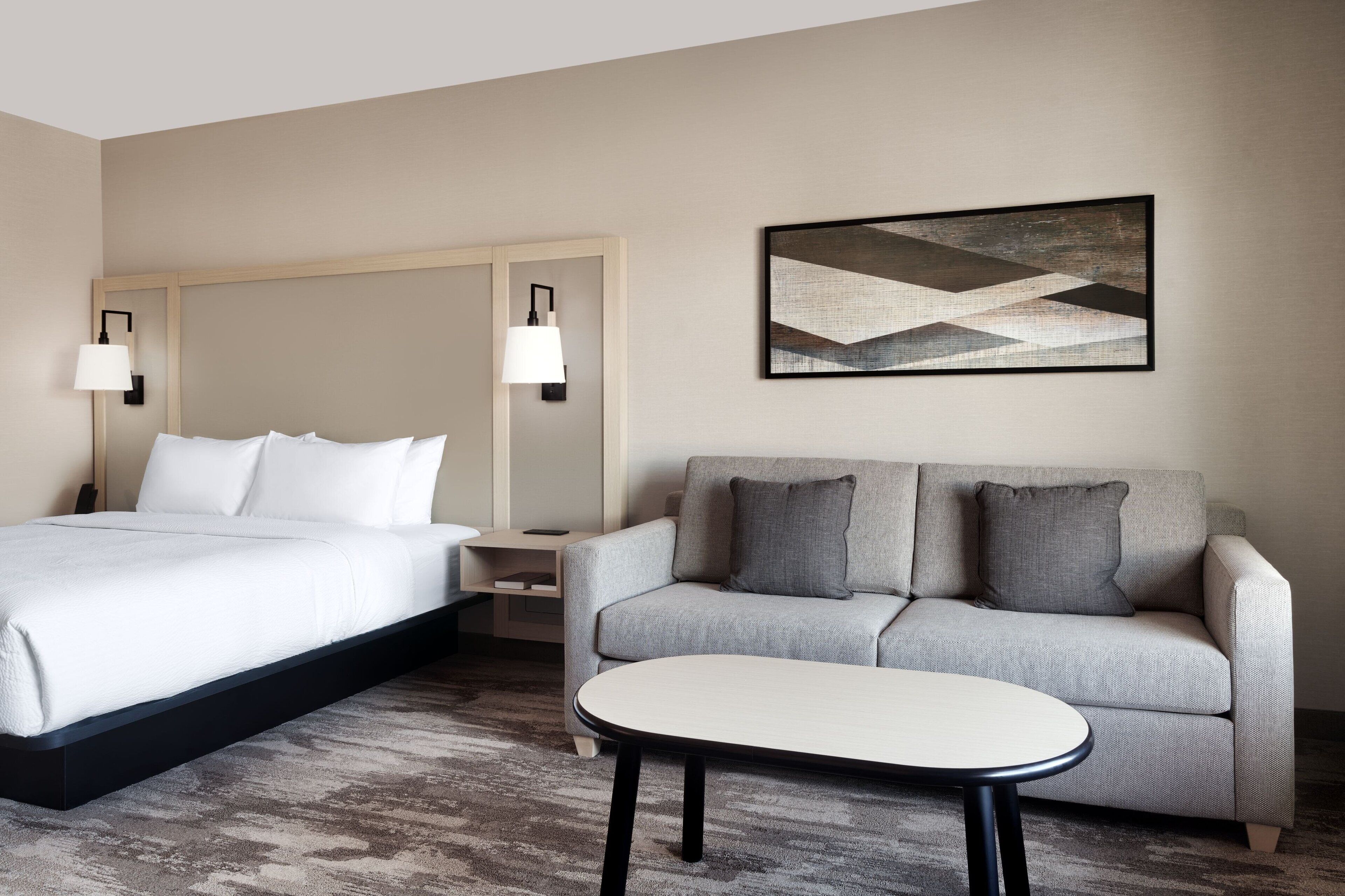 A hotel room with a bed, a sofa, a small table, and a wall-mounted lamp.