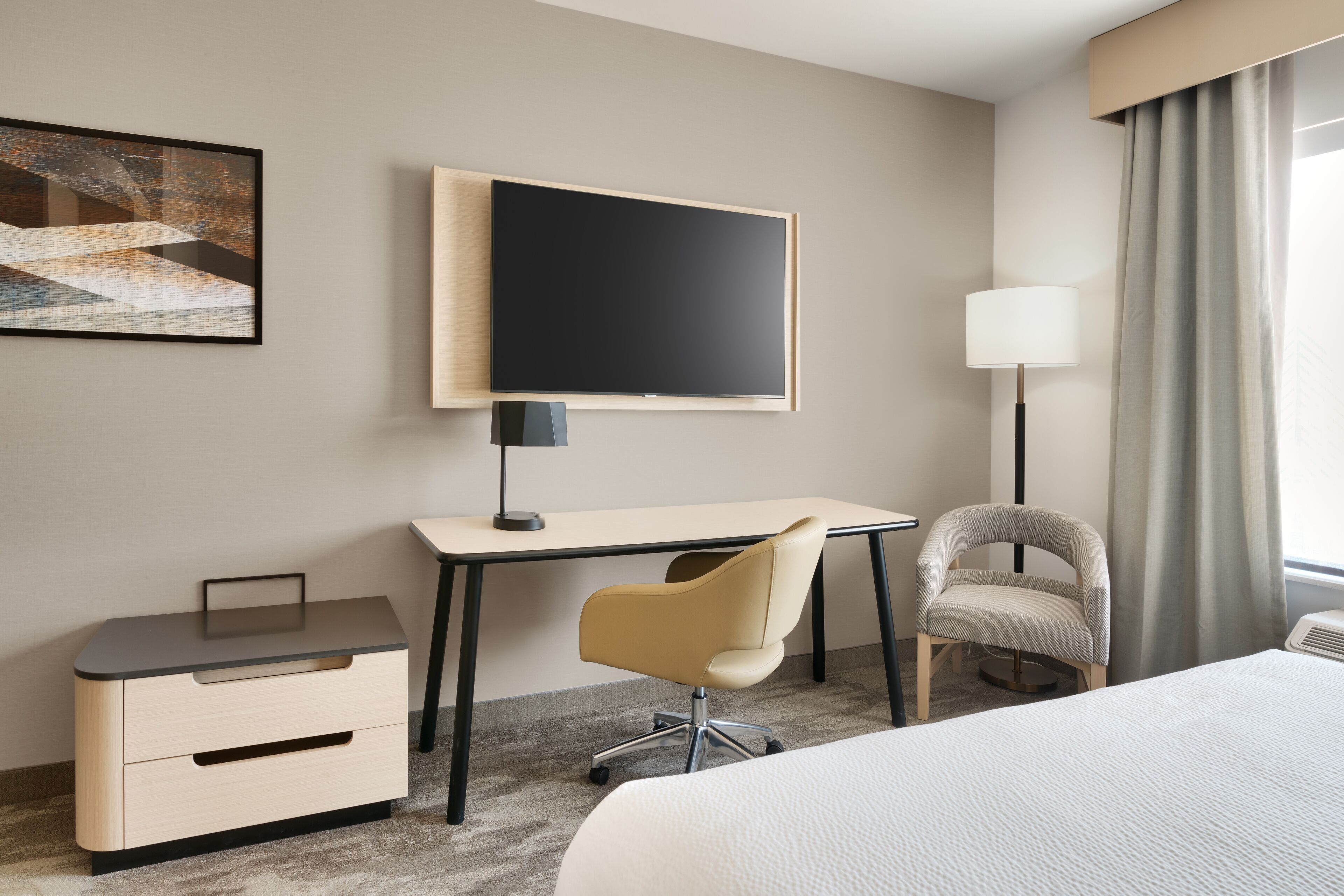 A hotel room with a bed, a desk with a chair, a television, a lamp, and a picture on the wall.