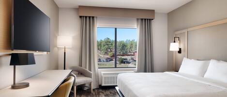 A hotel room with a bed, desk, chair, TV, and a window with a view of trees and parked cars.