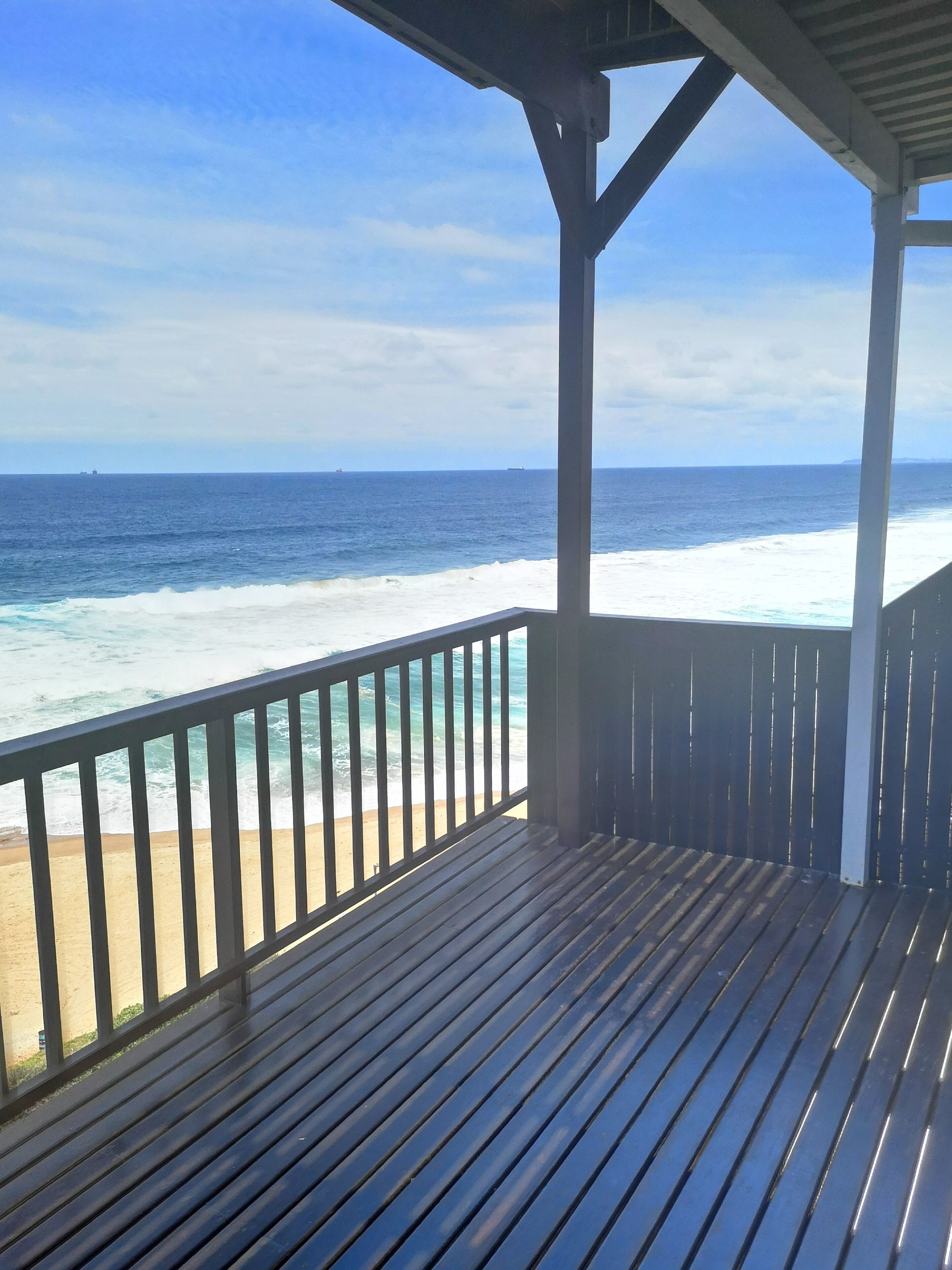 Une terrasse en bois avec une rambarde donnant sur une plage et l’océan.