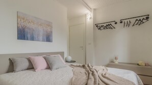 Et soveværelse med seng, puder, et tæppe, et kunstværk på væggen, en dør, en vægmonteret lampe, et lille bord med et stearinlys og en hylde med et stearinlys og blomster.