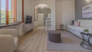 En moderne stue med sofa, skammel og et fjernsyn på et stativ. Der er et spiseområde med bord og stole, og et køkken i baggrunden.