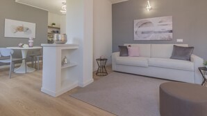 En moderne stue med en hvid sofa, et spiseområde med et hvidt bord og stole, og en grå væg med kunstværker.