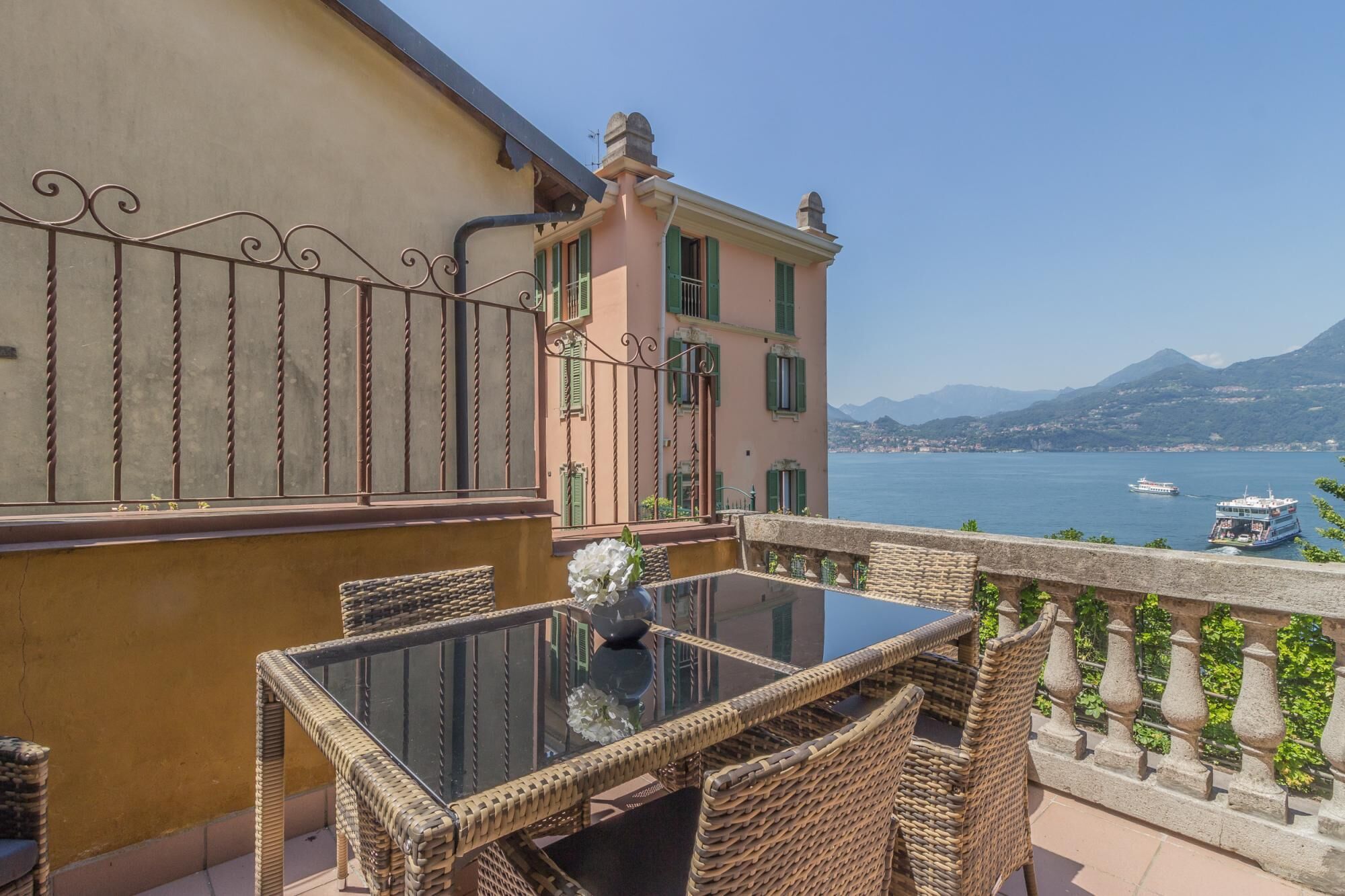 A balcony with wicker furniture, a glass table, and a view of the sea and mountains.