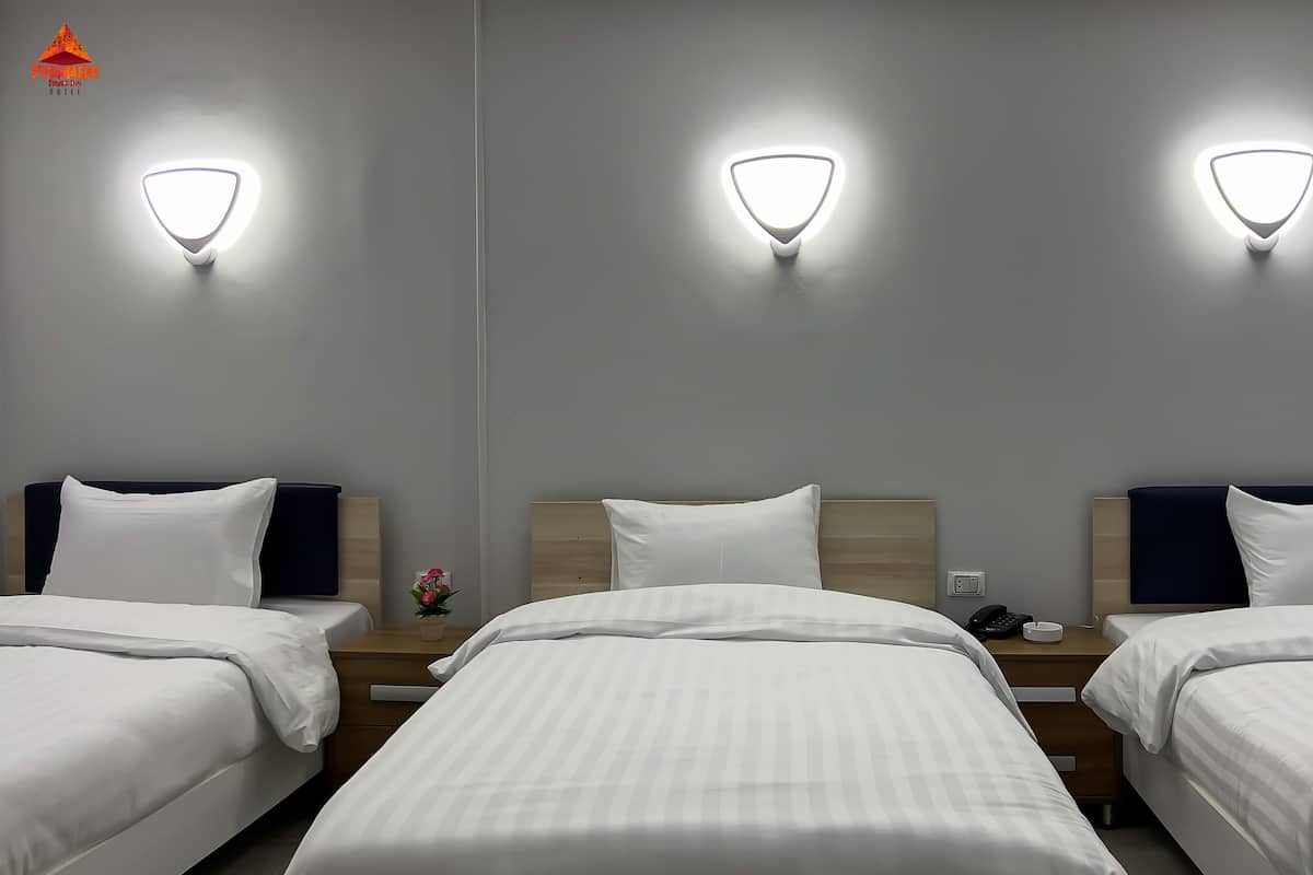 A hotel room with three beds, each with white bedding and headboards. There are wall-mounted lights above each bed.