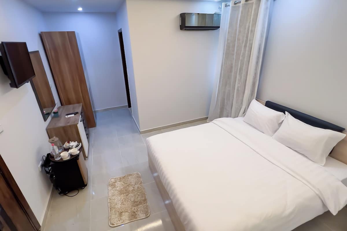 A hotel room with a bed, a desk with a chair, a flat-screen TV mounted on the wall, a small table with a lamp, and a window with curtains.