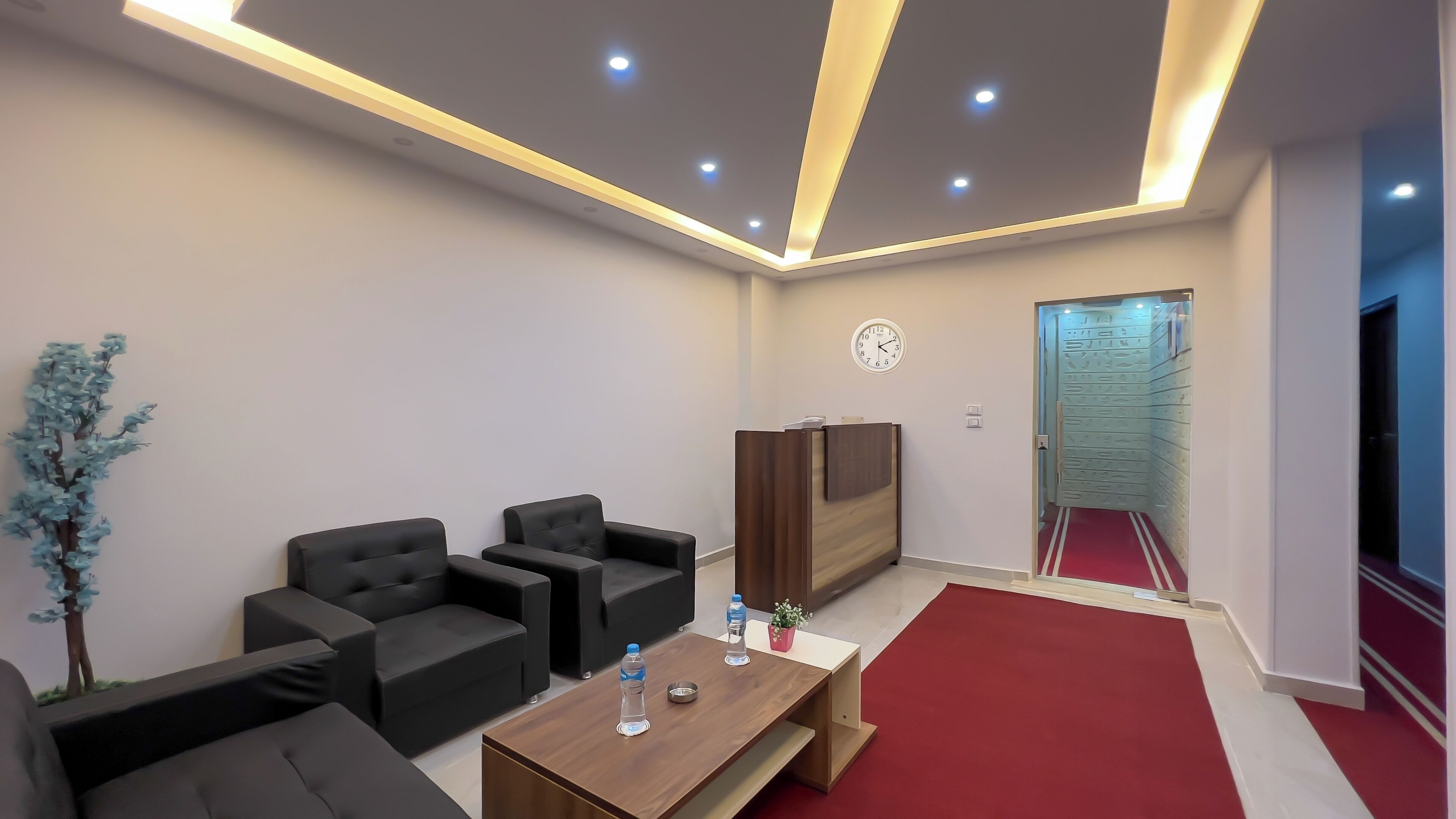 A modern waiting area with a reception desk, seating, a clock, and a red carpeted hallway.