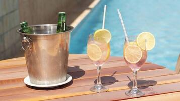 Cenário à beira da piscina com um balde de metal contendo garrafas, dois copos com rodelas de limão e uma mesa de madeira.