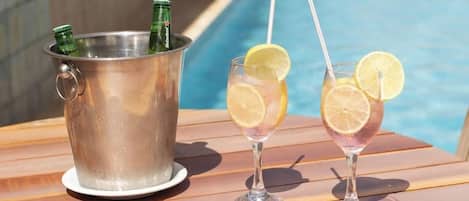Escena junto a la piscina con un cubo de metal que contiene botellas, dos vasos con rodajas de limón y una mesa de madera.