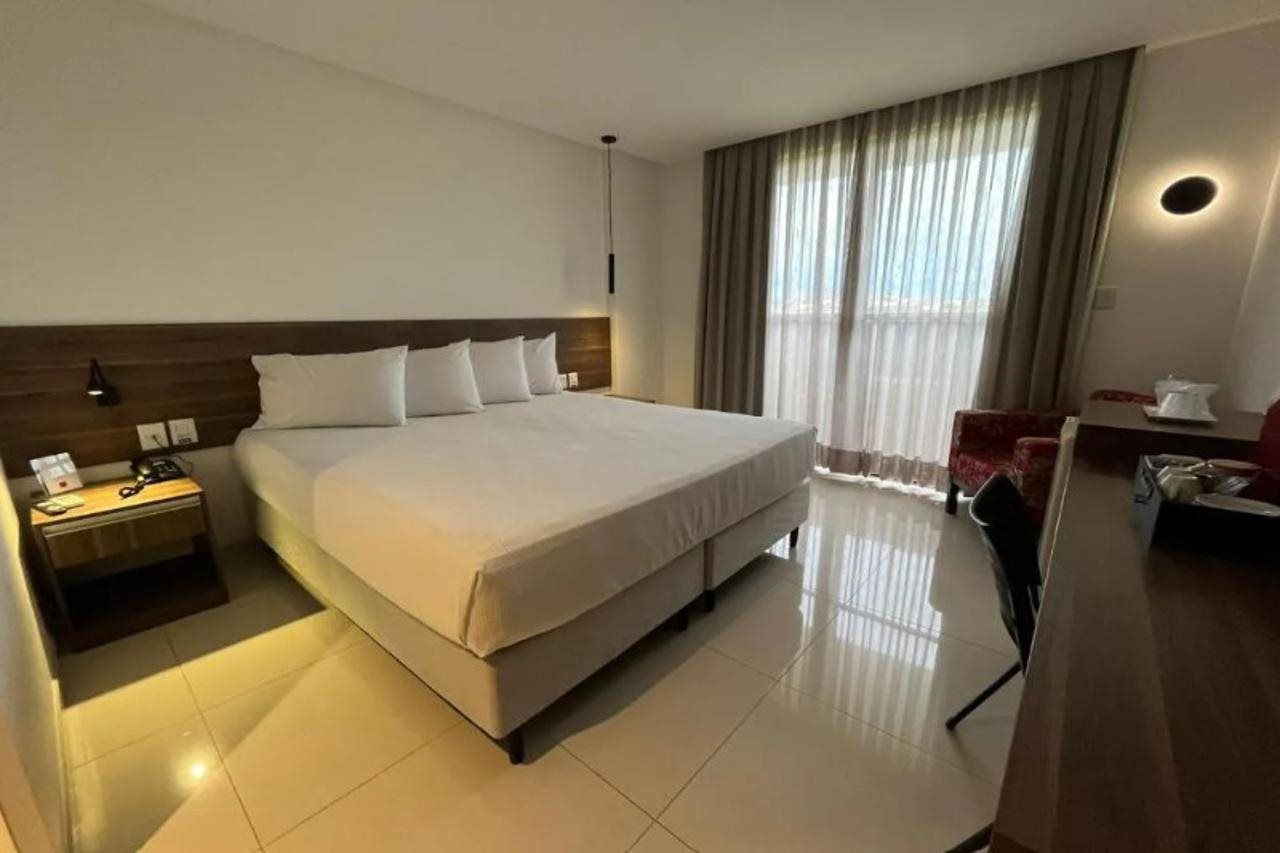 Quarto de hotel com uma cama grande, mesa de cabeceira com abajur, escrivaninha com cadeira e uma janela ampla com cortinas.
