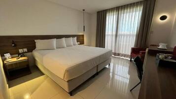 Quarto de hotel com uma cama grande, mesa de cabeceira com abajur, escrivaninha com cadeira e uma janela ampla com cortinas.