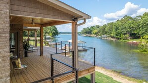 Una terraza de madera con mobiliario exterior, un espacio cubierto y vistas a un lago con barcos.