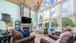Un salón amplio con sofás de cuero, una chimenea, un televisor de pantalla plana y ventanas grandes que ofrecen vistas al exterior.