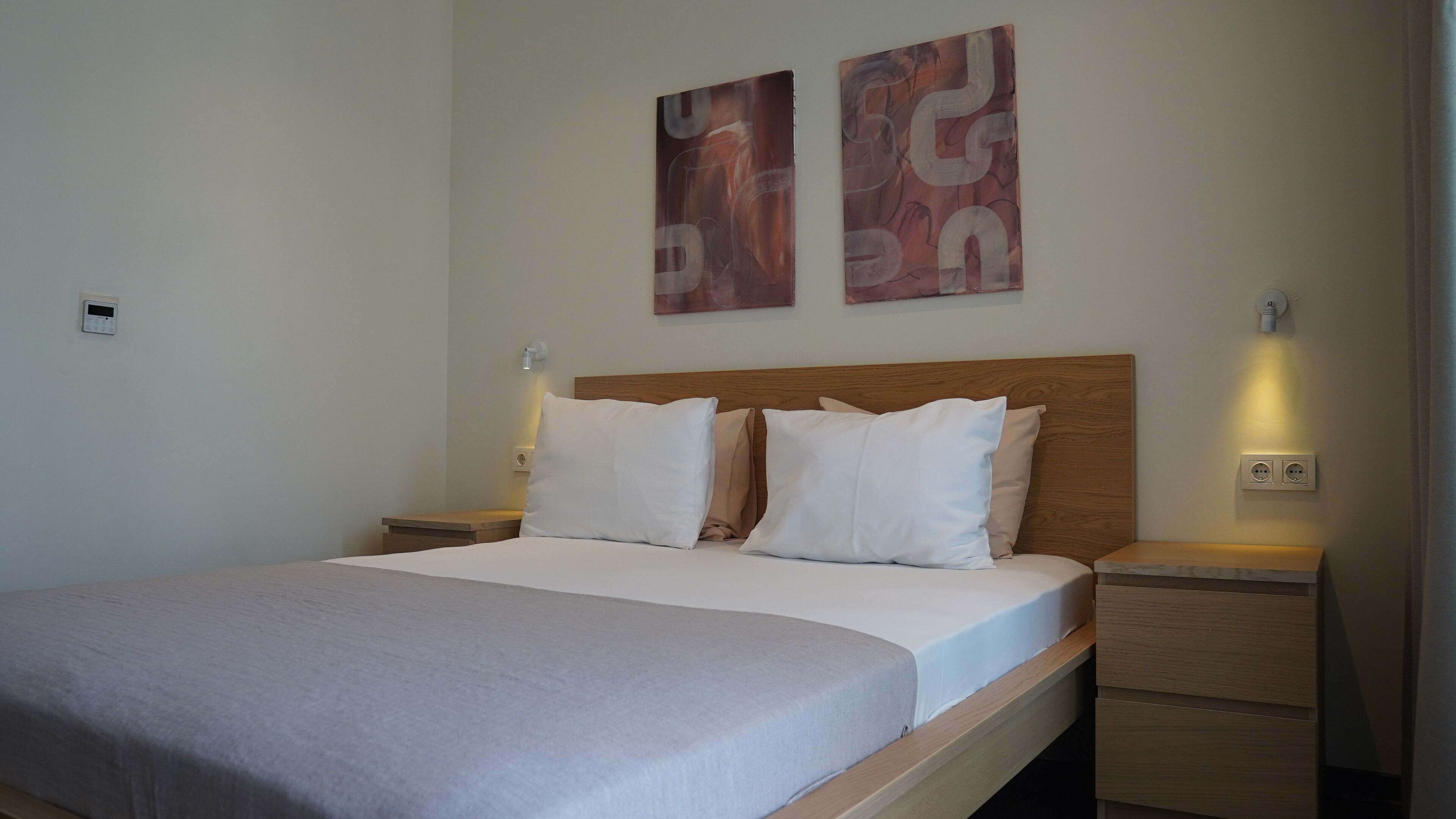 Un lit bien fait, avec du linge de lit blanc et une tête de lit en bois. Il y a deux tables de chevet, chacune avec une lampe. Les murs sont de couleur unie et ornés de deux tableaux abstraits.