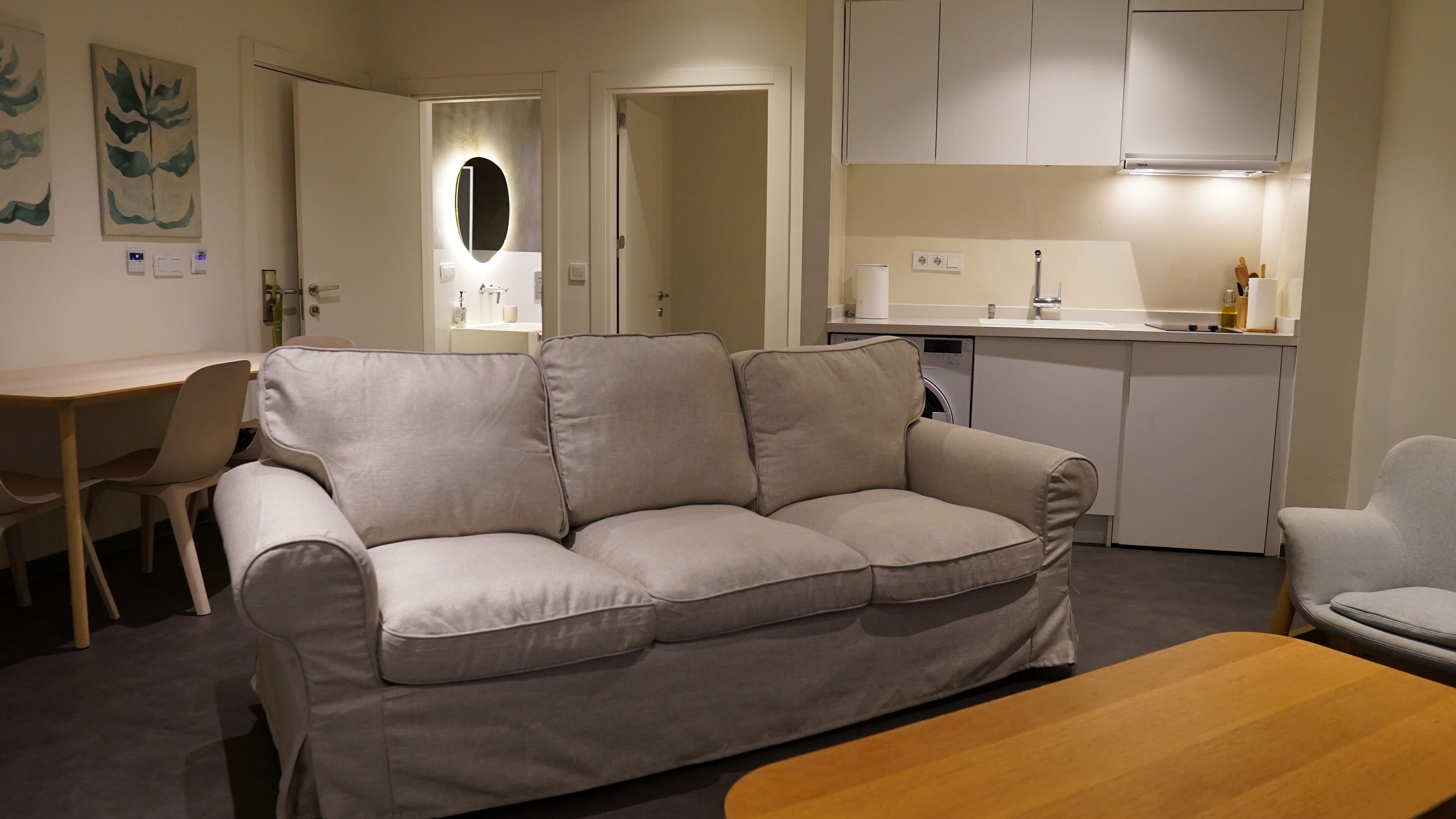 A living room with a sofa, a wooden coffee table, a dining table with chairs, a kitchen area with cabinets and a sink, and a hallway with a door.