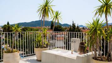 A rooftop terrace with a hot tub, potted plants, and a view of a cityscape and mountains.