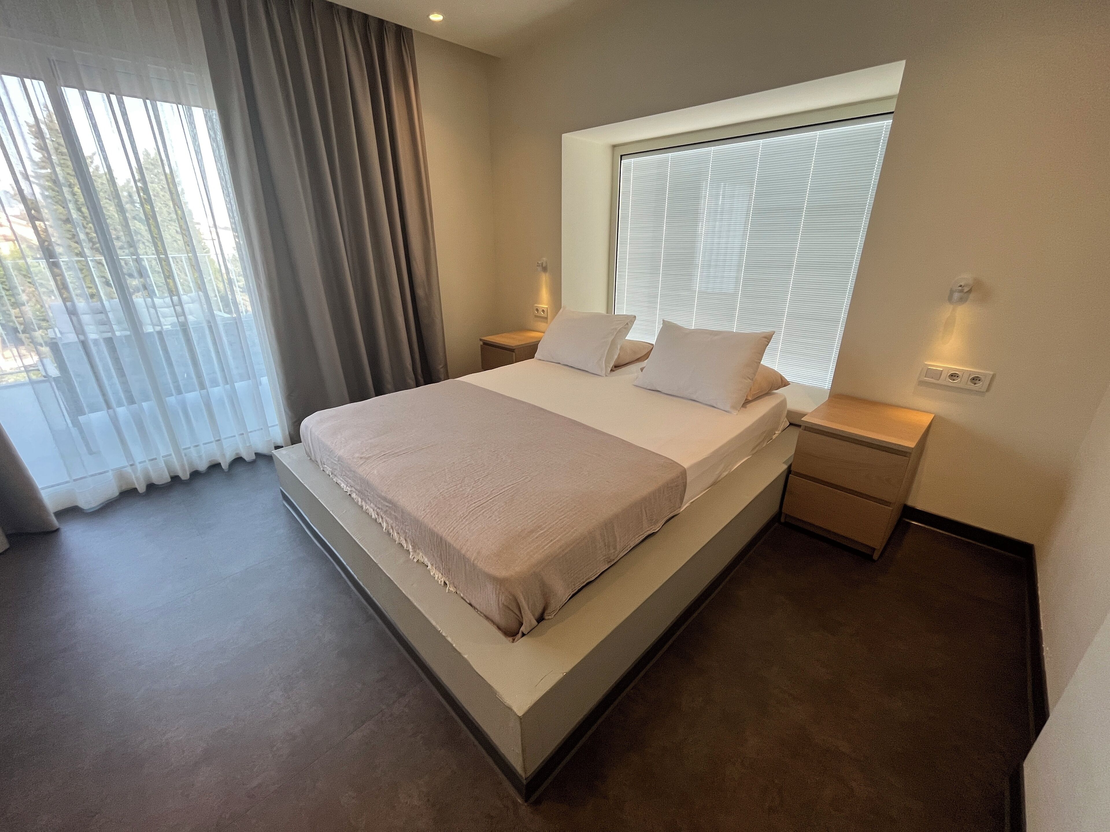 A modern bedroom with a large bed, a window with blinds, a bedside table, and a nightstand.