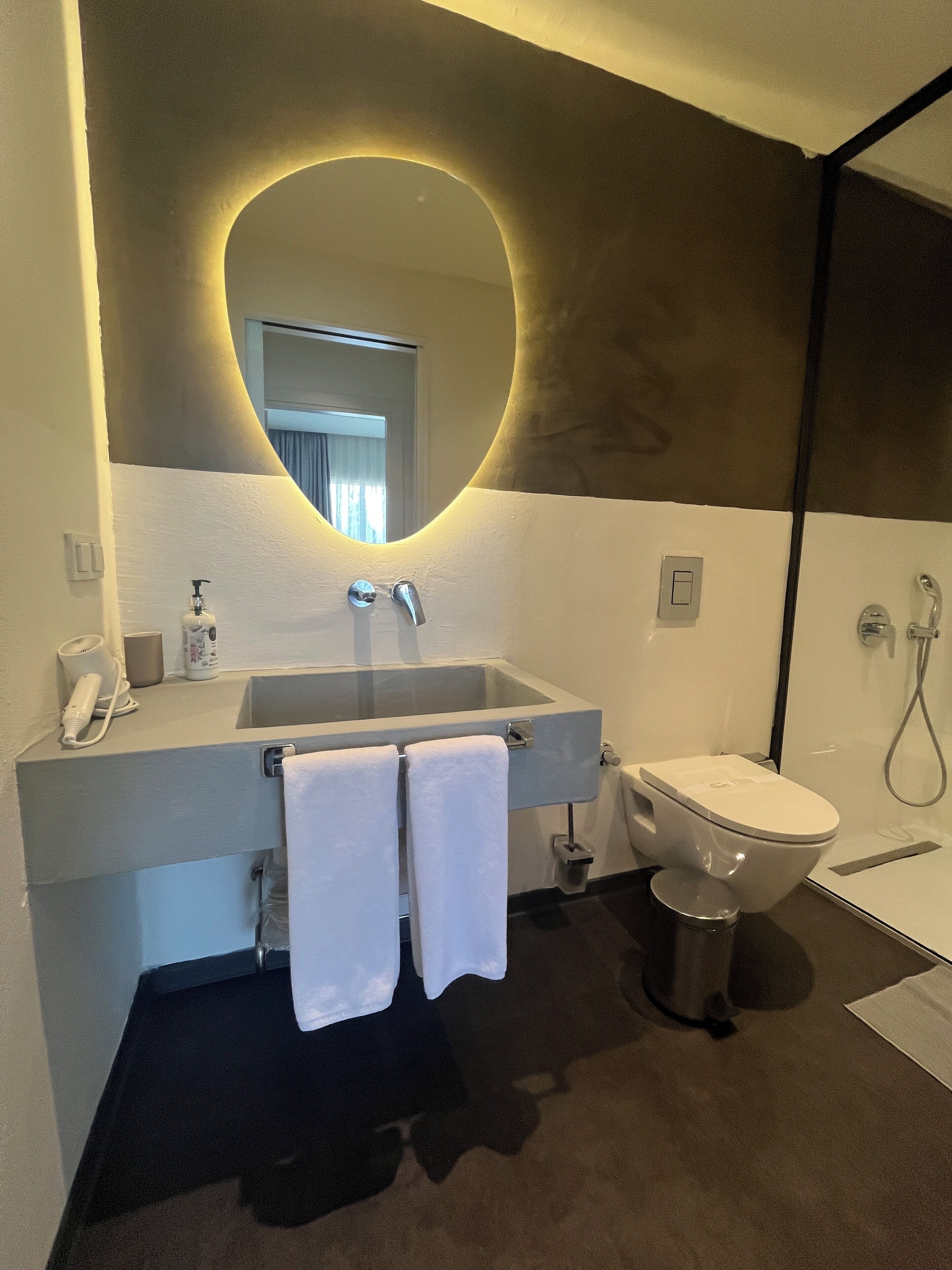 A modern bathroom with a large oval mirror, a sink with a faucet, a toilet, and a shower area.