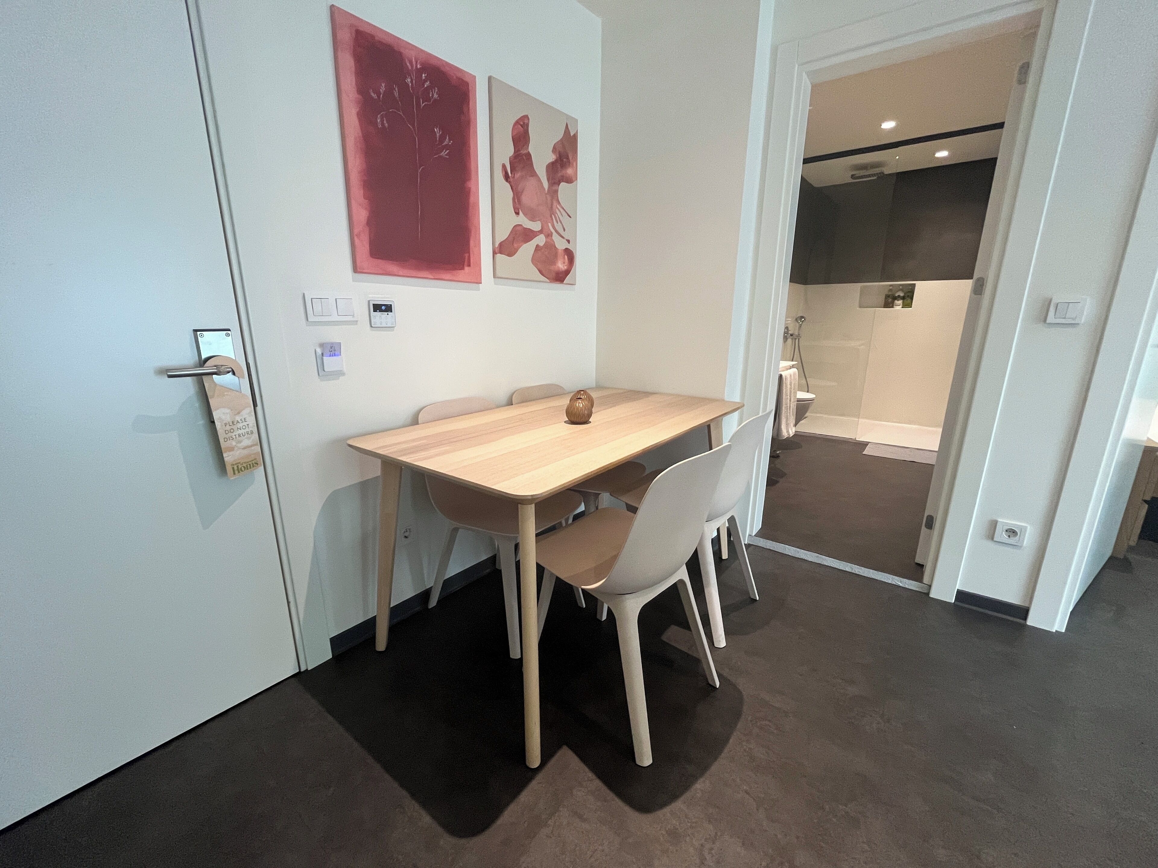 A modern hotel room with a wooden dining table, white chairs, and a small vase. There are two paintings on the wall and a door with a 'Do Not Disturb' sign.