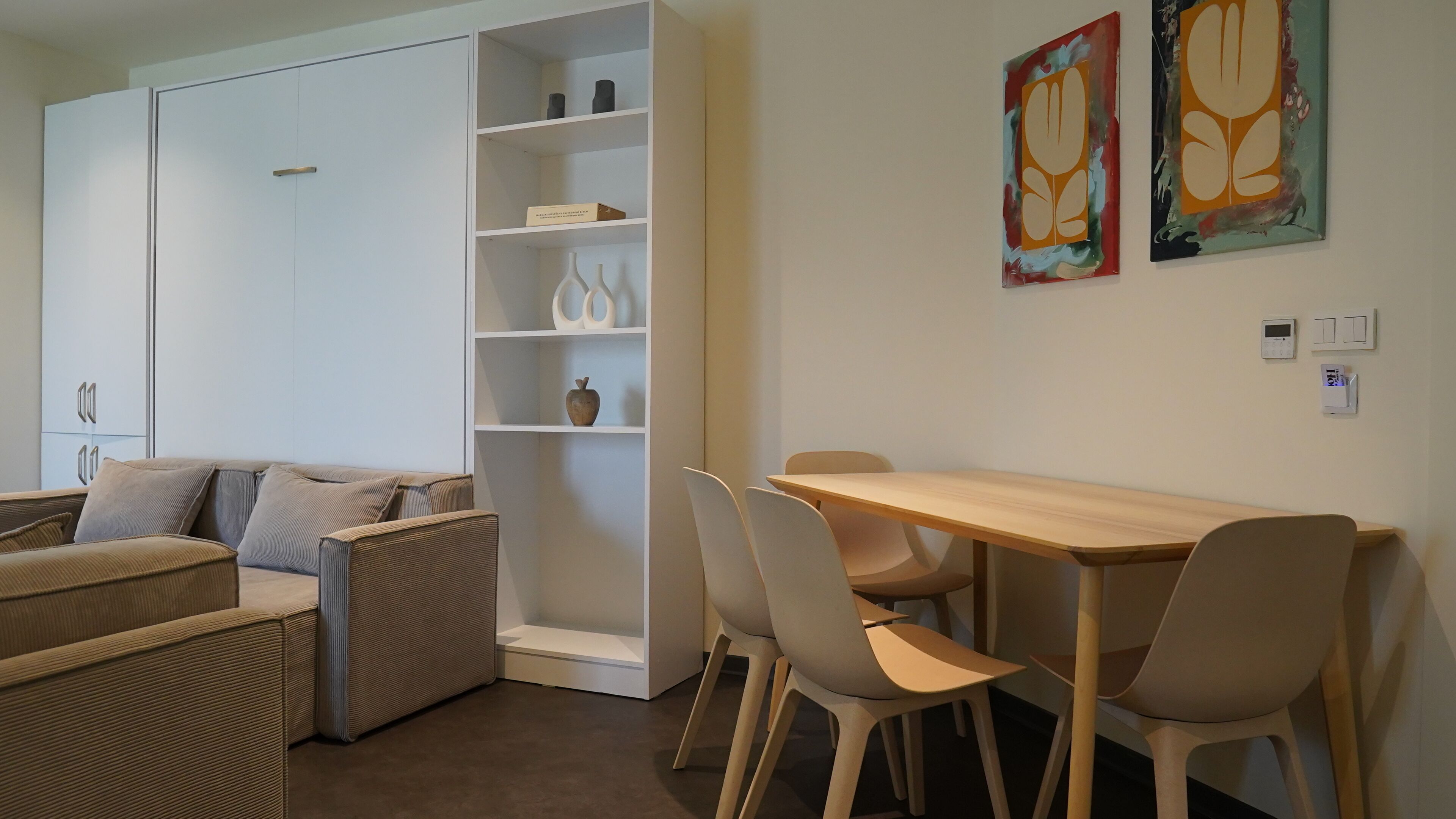 A modern living room with a sofa, a wooden table, and chairs. There is a white cabinet with shelves and a small table. Two abstract paintings are on the wall.