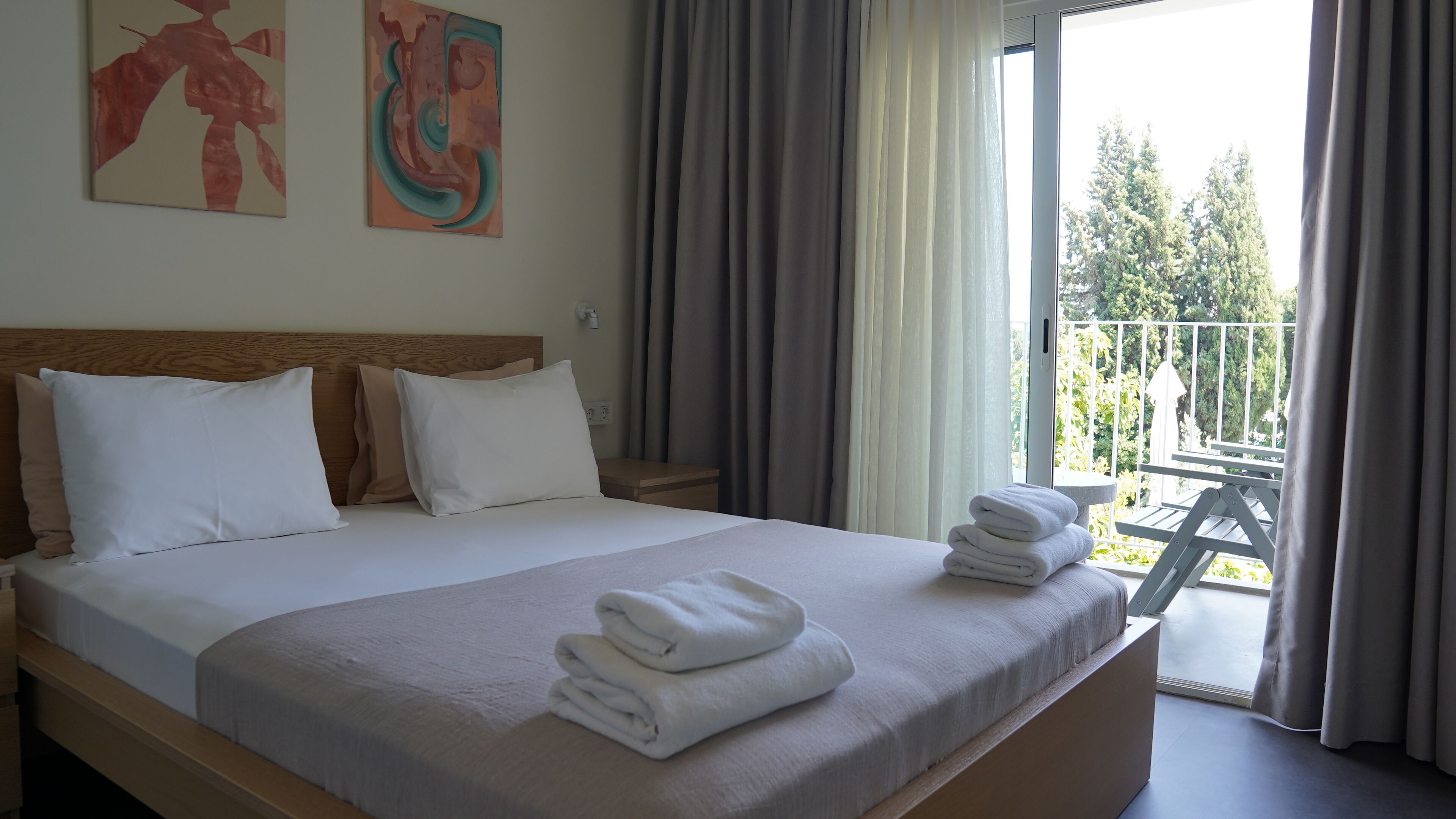 A hotel room with a large bed, a wooden headboard, and a balcony with a view of trees.