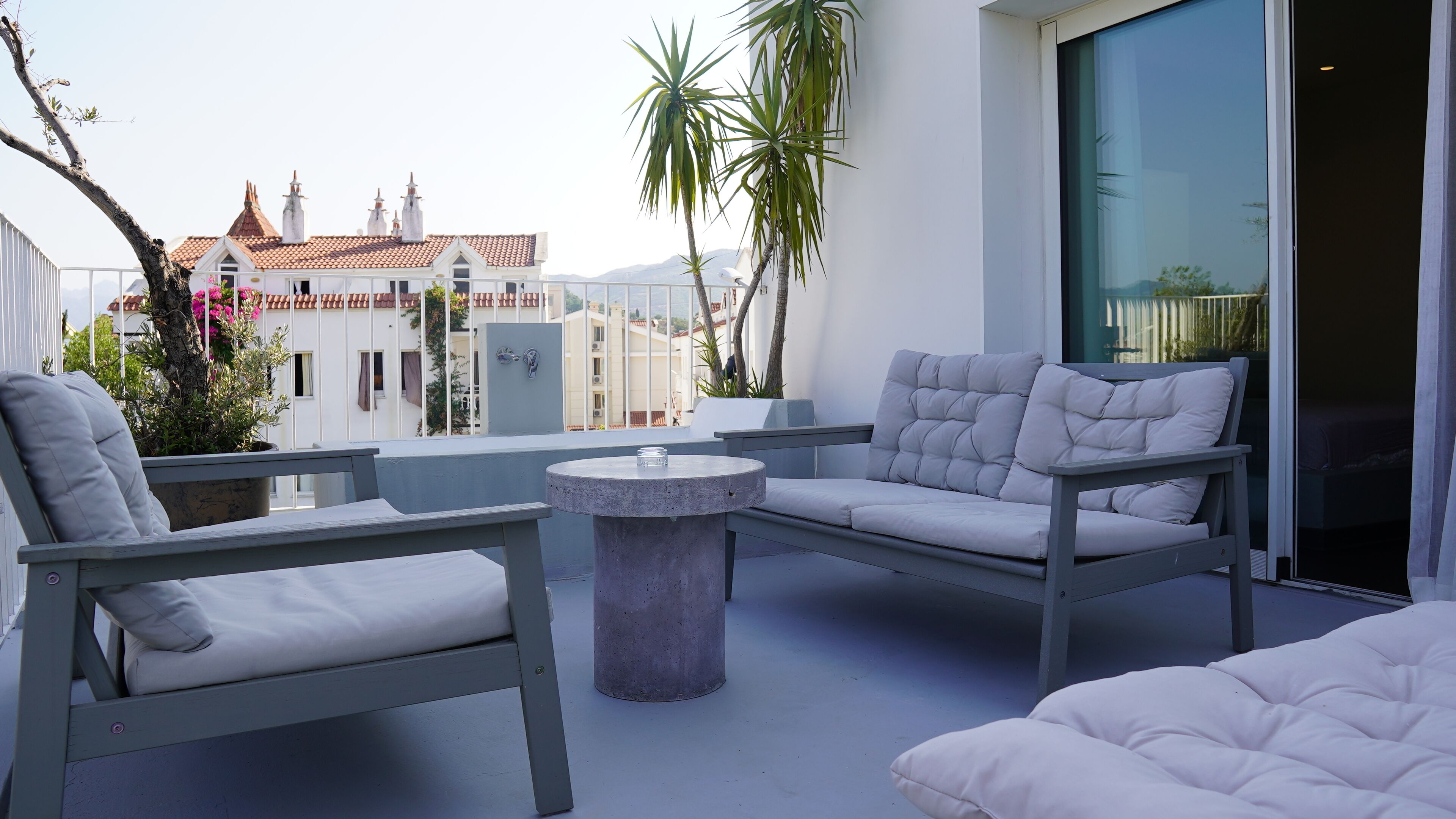 En balkon med siddeplads, herunder to stole og et bord, med udsigt over bybilledet med bygninger og en klar himmel.