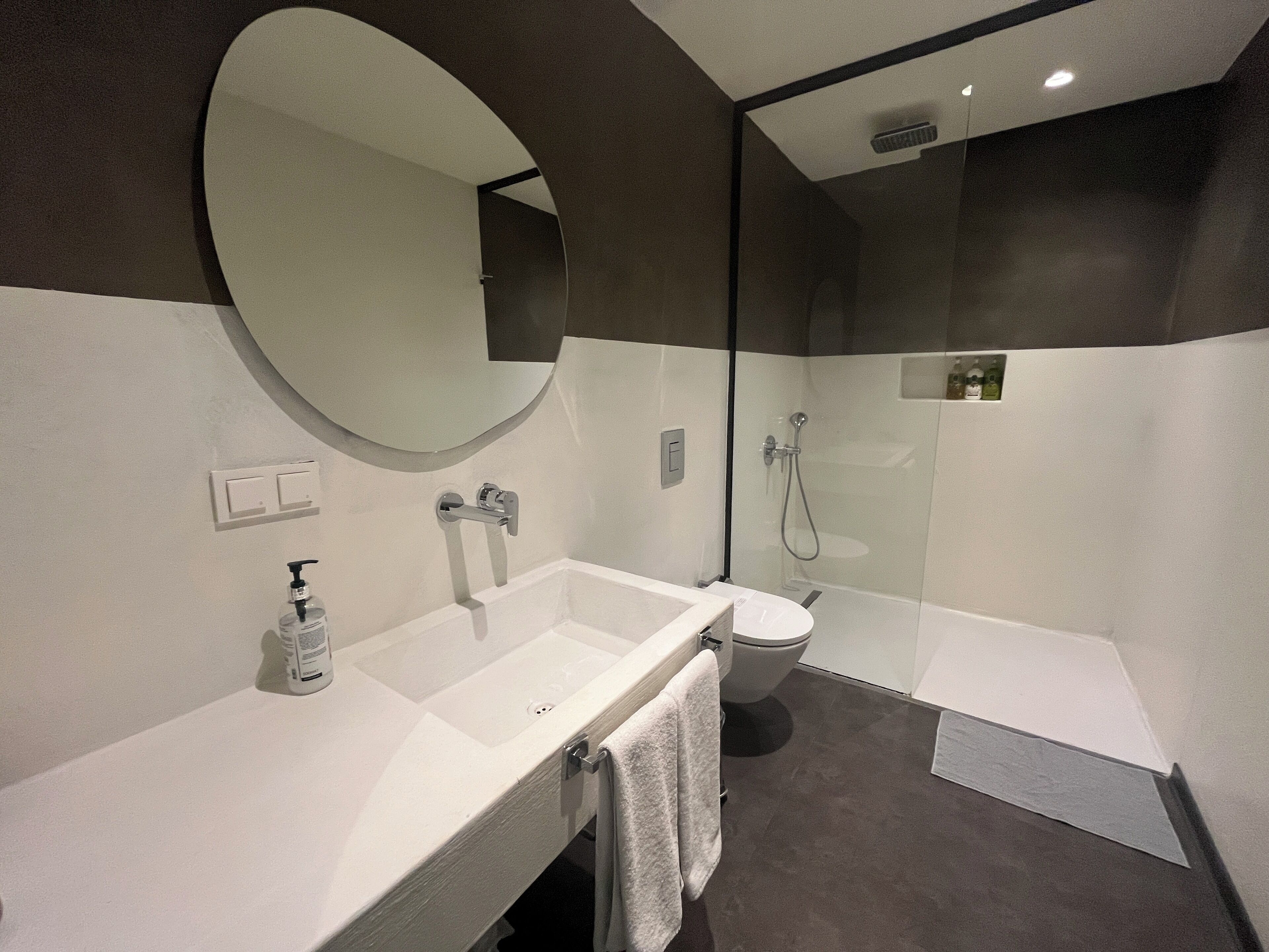 Un baño moderno con un espejo ovalado grande, un lavamanos blanco, un inodoro y una zona de ducha con mampara de vidrio.