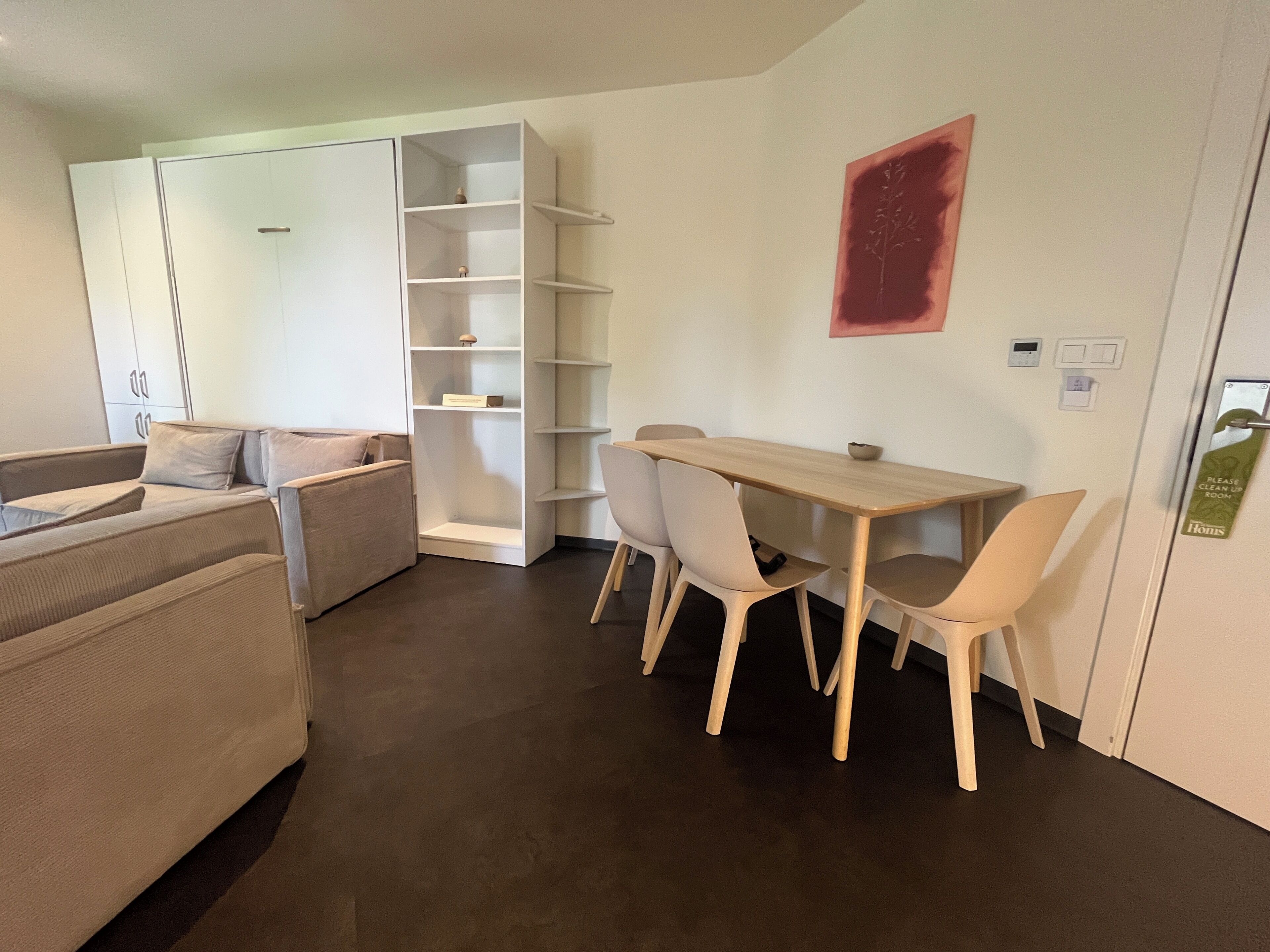 A modern hotel room with a sofa, a dining table with chairs, a white shelving unit, and a red abstract painting on the wall.