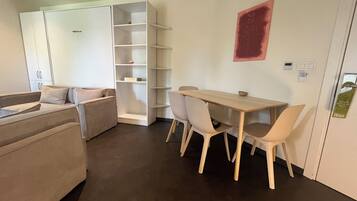 A modern hotel room with a sofa, a dining table with chairs, a white shelving unit, and a red abstract painting on the wall.