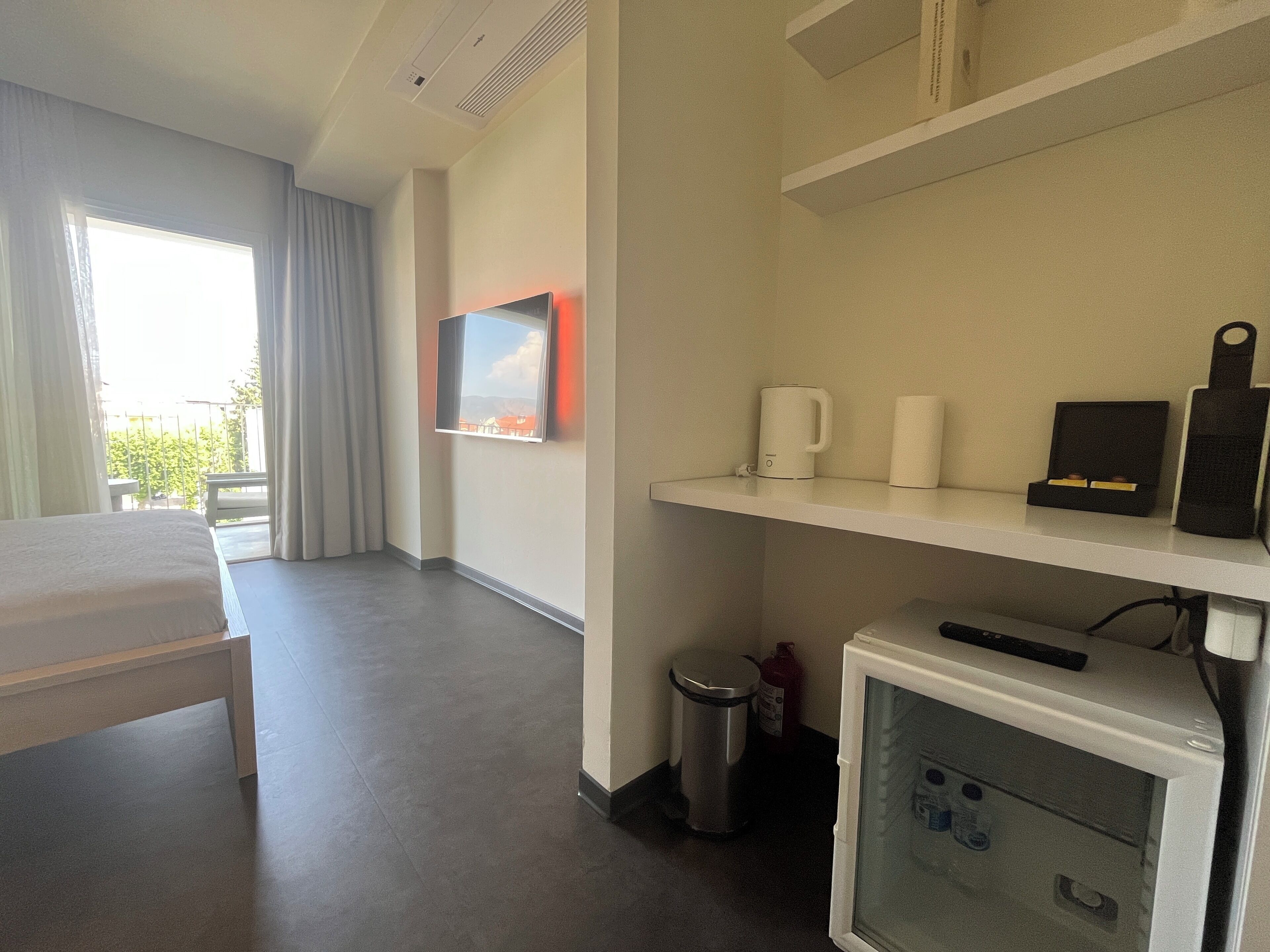 A hotel room with a bed, a TV mounted on the wall, a small shelf with a coffee maker, a trash bin, and a refrigerator.