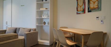 Una sala de estar moderna con un sofá, una mesa de comedor con sillas, un estante blanco con objetos decorativos y dos cuadros abstractos en la pared.