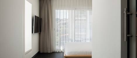Una habitación de hotel moderna con una cama, un televisor montado en la pared y un gran ventanal con cortinas.